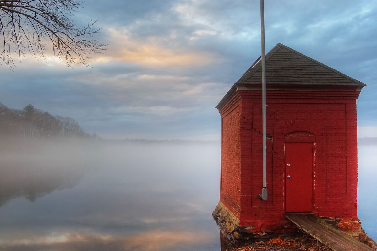 A red brick pump house with a flagpole stands at the edge of a mist-covered lake under soft morning clouds.