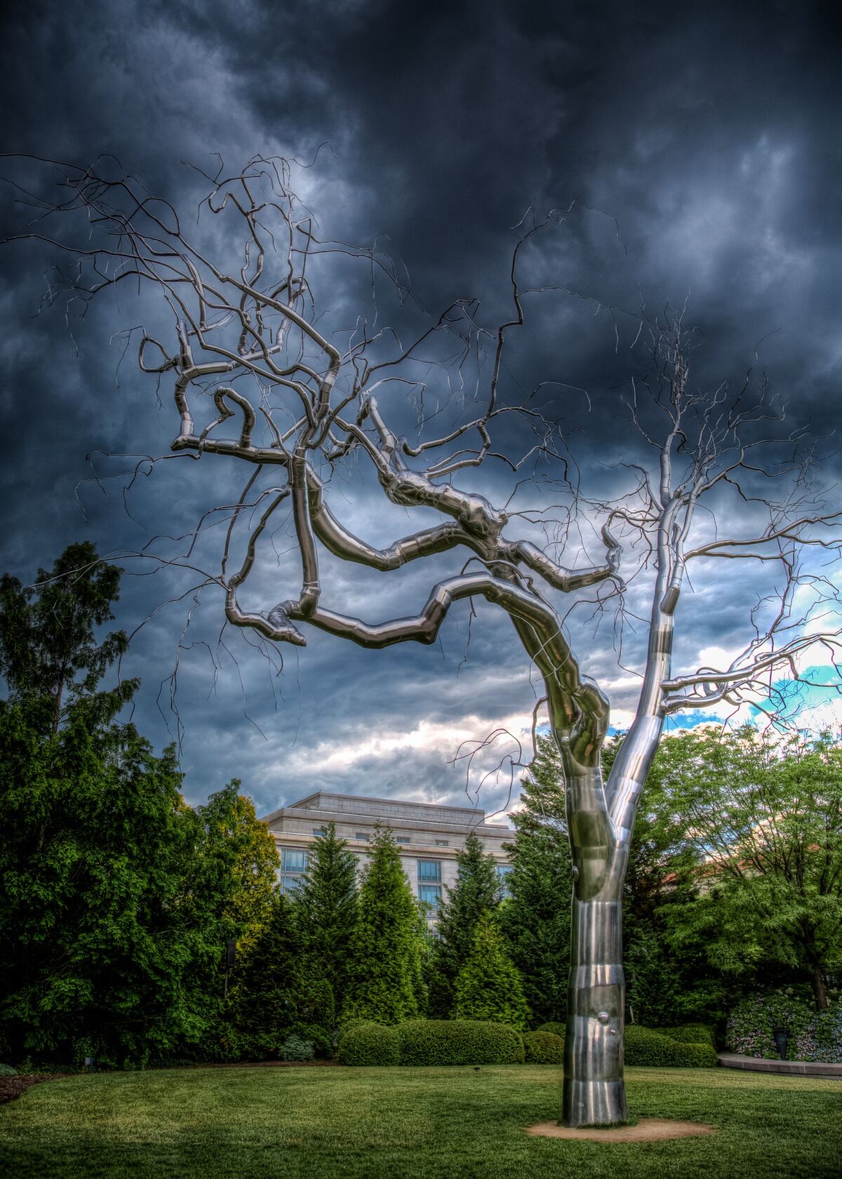 A leafless stainless-steel tree sculpture rises from a green lawn against dark storm clouds.