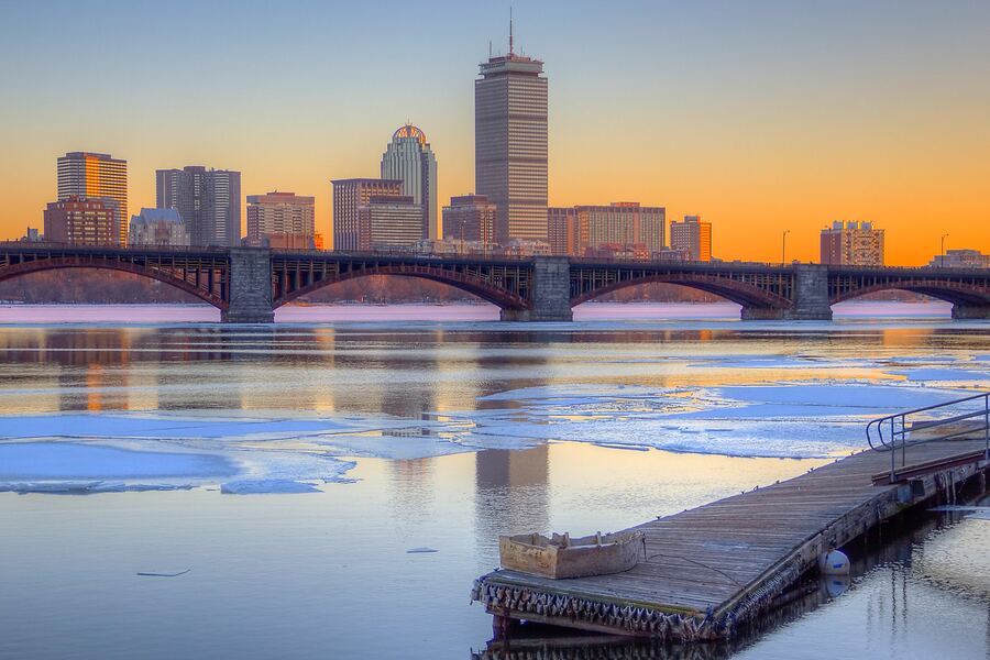 The Boston skyline at sunset beyond the Longfellow Bridge, with a wooden dock in the foreground and ice floes on the Charles River.