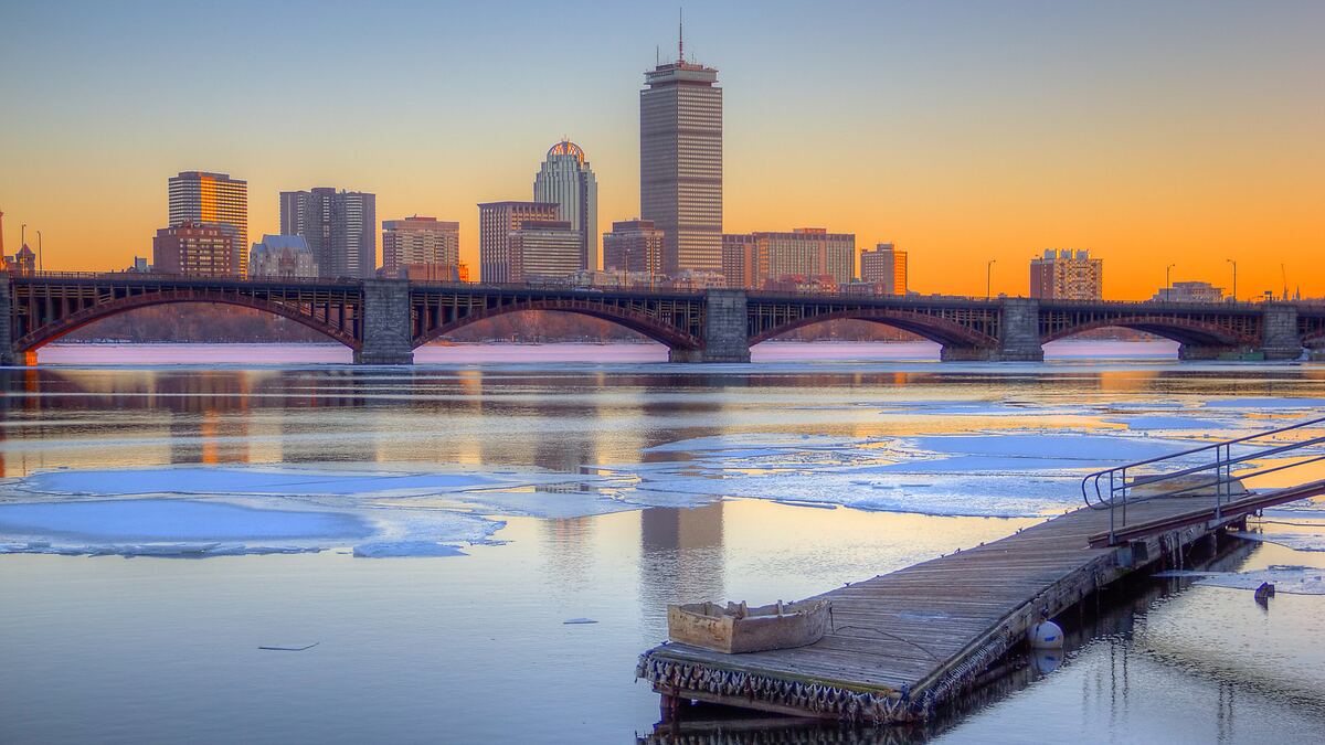 The Boston skyline at sunset beyond the Longfellow Bridge, with a wooden dock in the foreground and ice floes on the Charles River.