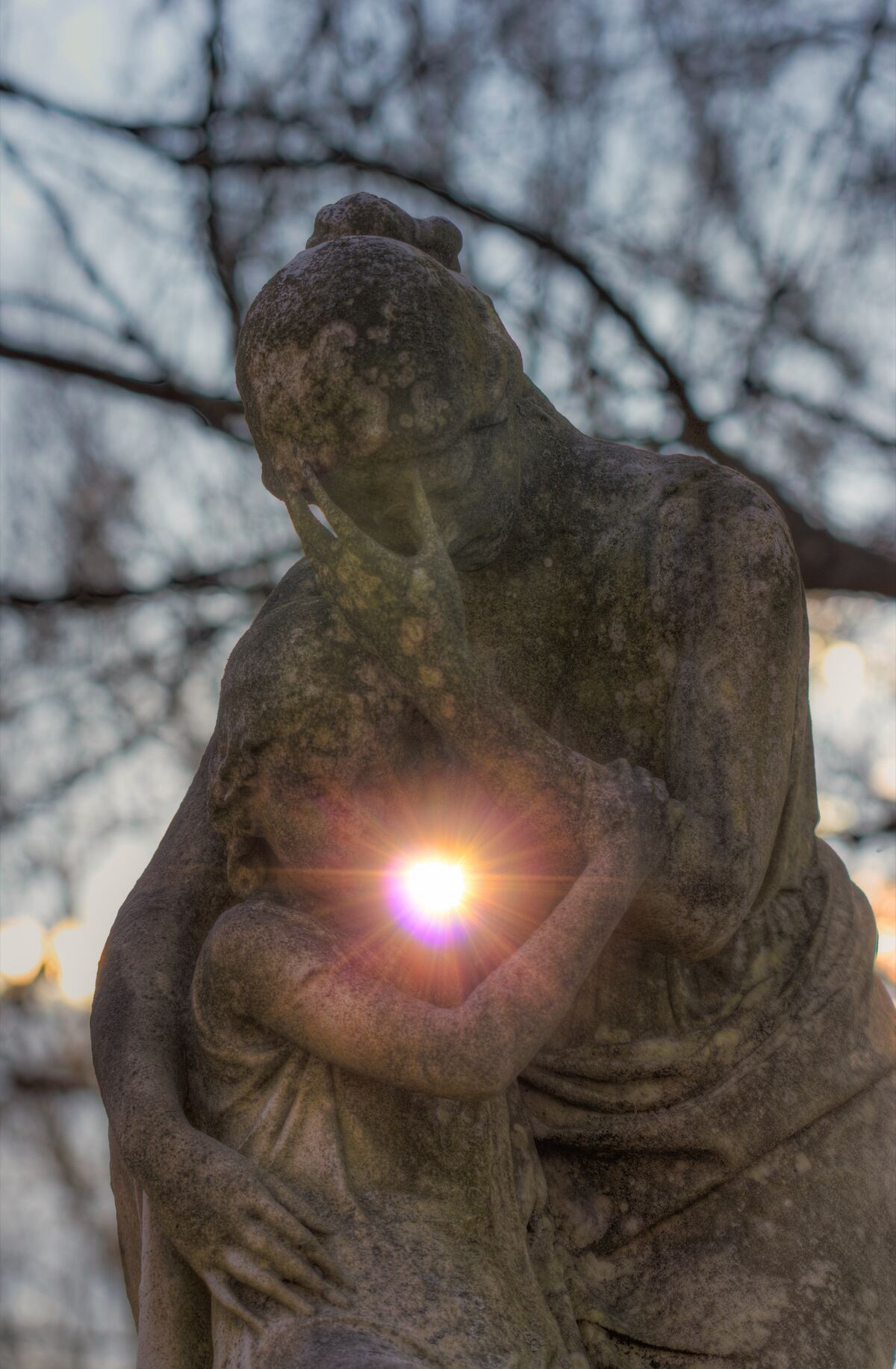 A weathered stone statue of a woman bending over a child, with the sun flaring between the two figures and bare branches behind.