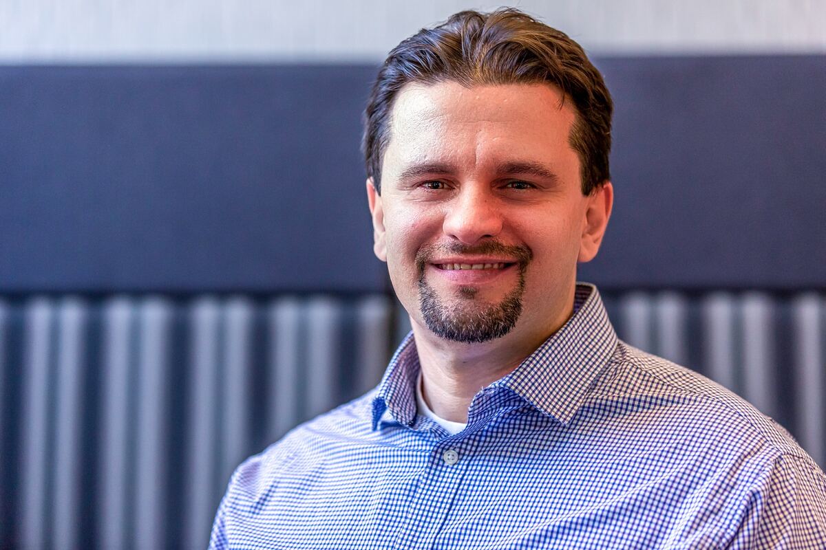 A man with short brown hair and a goatee smiles in a blue checked shirt against a blurred gray-paneled wall.