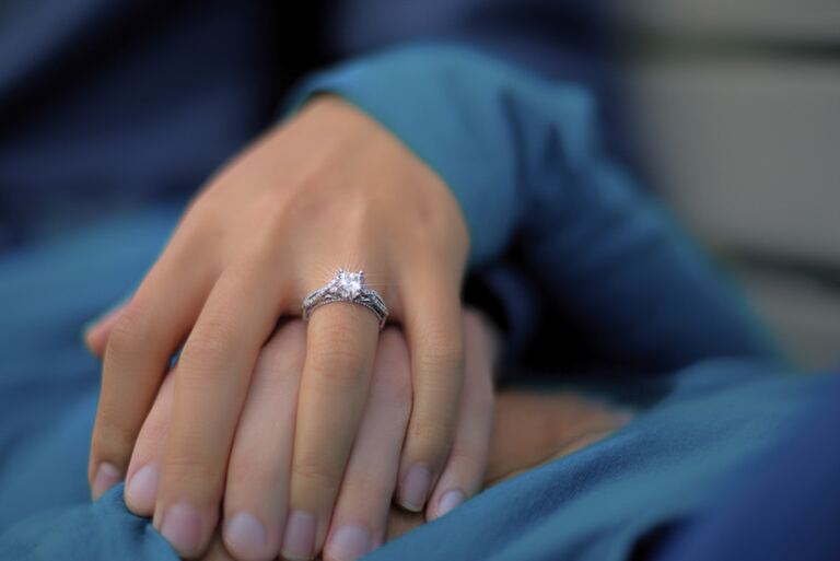A diamond engagement ring sparkles on a woman's hand resting atop another hand, framed by teal-blue clothing.