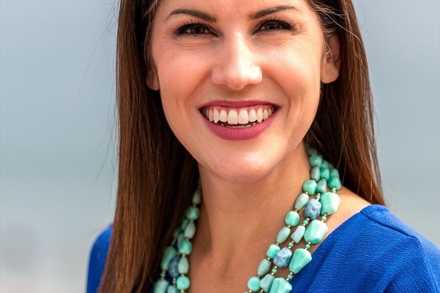 A smiling woman with long brown hair wears a royal-blue top and a layered turquoise bead necklace against a soft, hazy ocean backdrop.