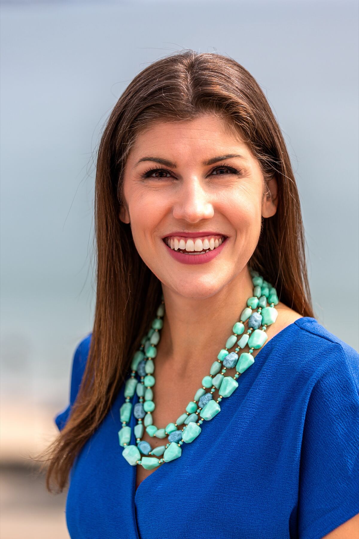 A smiling woman with long brown hair wears a royal-blue top and a layered turquoise bead necklace against a soft, hazy ocean backdrop.