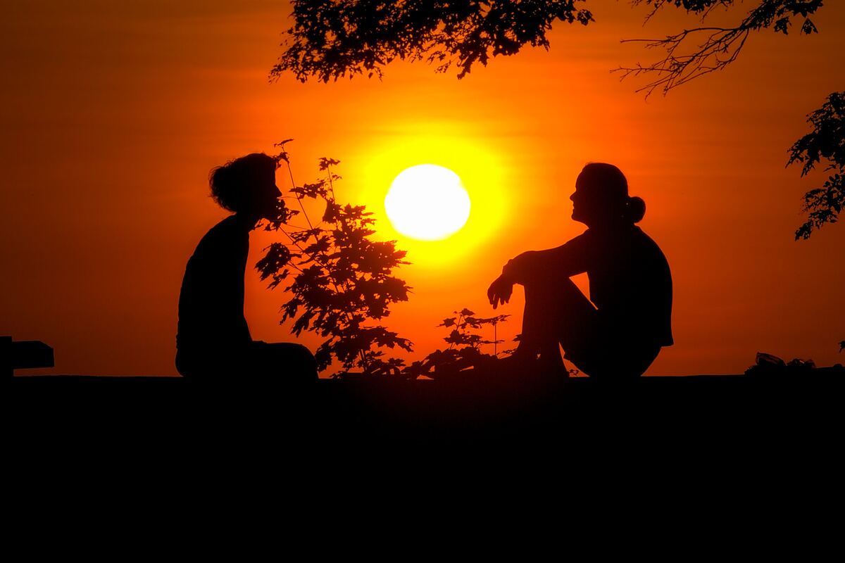 Two people sit facing each other in silhouette on a wall at sunset, the sun glowing orange between them beneath overhanging branches.